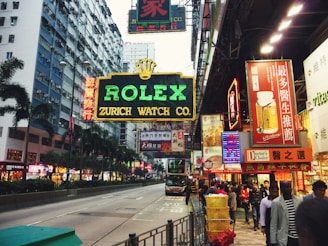 A bustling urban street scene with a prominent neon sign advertising Rolex Zurich Watch Co. Buildings line the street, adorned with a plethora of vibrant signs and advertisements in various languages, including Chinese characters. The atmosphere is lively, with many people walking along the sidewalk, creating a sense of busy city life. Palm trees are visible along the median, and a double-decker bus is approaching in the background.