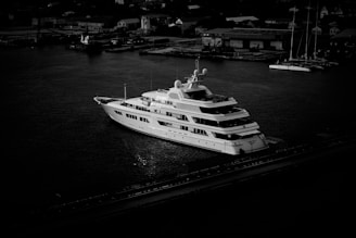 A large, luxurious yacht is anchored in a harbor, surrounded by calm water. Several small buildings and structures are visible in the background, suggesting an industrial or urban setting. The image is in black and white, casting dramatic shadows and highlighting the yacht's sleek design.