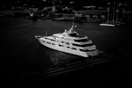 A large, luxurious yacht is anchored in a harbor, surrounded by calm water. Several small buildings and structures are visible in the background, suggesting an industrial or urban setting. The image is in black and white, casting dramatic shadows and highlighting the yacht's sleek design.