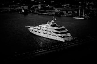 A large, luxurious yacht is anchored in a harbor, surrounded by calm water. Several small buildings and structures are visible in the background, suggesting an industrial or urban setting. The image is in black and white, casting dramatic shadows and highlighting the yacht's sleek design.