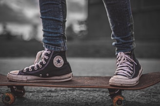 A close-up view of a person wearing black high-top sneakers standing on a skateboard. The sneakers are scuffed and well-worn, with white laces and a prominent logo on the side. The skateboard is on a paved surface, and the person's jeans are rolled up slightly at the ankles. The background is blurred, highlighting the focus on the footwear and skateboard.
