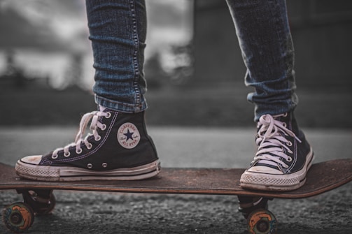 A close-up view of a person wearing black high-top sneakers standing on a skateboard. The sneakers are scuffed and well-worn, with white laces and a prominent logo on the side. The skateboard is on a paved surface, and the person's jeans are rolled up slightly at the ankles. The background is blurred, highlighting the focus on the footwear and skateboard.