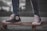 A close-up view of a person wearing black high-top sneakers standing on a skateboard. The sneakers are scuffed and well-worn, with white laces and a prominent logo on the side. The skateboard is on a paved surface, and the person's jeans are rolled up slightly at the ankles. The background is blurred, highlighting the focus on the footwear and skateboard.