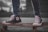 A close-up view of a person wearing black high-top sneakers standing on a skateboard. The sneakers are scuffed and well-worn, with white laces and a prominent logo on the side. The skateboard is on a paved surface, and the person's jeans are rolled up slightly at the ankles. The background is blurred, highlighting the focus on the footwear and skateboard.