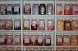 A collection of jars filled with a variety of colorful candies is arranged neatly on white shelves. Each jar has a pink label with the name of the candies inside, such as cola bottles, fizzy snakes, and tongue painters. The candies are in different shapes and colors, ranging from red and yellow gummy worms to white and pink fizzy sweets.