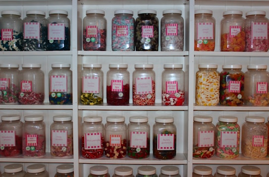 A collection of jars filled with a variety of colorful candies is arranged neatly on white shelves. Each jar has a pink label with the name of the candies inside, such as cola bottles, fizzy snakes, and tongue painters. The candies are in different shapes and colors, ranging from red and yellow gummy worms to white and pink fizzy sweets.
