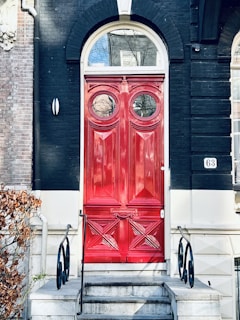 A bold red door with intricate decorative panels is set within a dark brick building. The door features two round windows at the top. Stone steps with black wrought iron railings lead up to it, and there is a small number plaque on the right side of the doorframe.