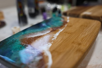 A wooden board with a decorative resin art design featuring an ocean-like pattern. The art includes gradients of teal, blue, and white, mimicking waves and sea foam on the shore. The background is blurred, emphasizing the focus on the board's intricate design.