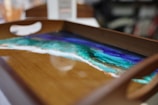 A wooden tray with a resin art design featuring a vibrant seascape pattern in shades of blue, green, and white. The colors appear to blend seamlessly, resembling an ocean scene with a sandy shore.