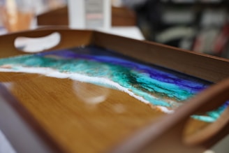 A wooden tray with a resin art design featuring a vibrant seascape pattern in shades of blue, green, and white. The colors appear to blend seamlessly, resembling an ocean scene with a sandy shore.