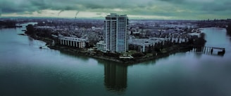 Tall residential building on a small piece of land surrounded by water, with adjacent lower buildings and a sprawling cityscape in the background under a cloudy sky.