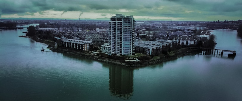 Tall residential building on a small piece of land surrounded by water, with adjacent lower buildings and a sprawling cityscape in the background under a cloudy sky.