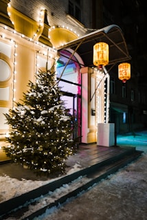 An outdoor scene features a decorated pine tree covered in snow, illuminated by white string lights. Brightly lit, ornate lanterns hang from an overhead awning in front of a warmly lit building entrance, creating a festive atmosphere. Snow is lightly scattered on the ground and steps leading up to the entrance.