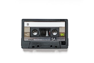 A black audio cassette tape with a side labeled 'B' and marked with 'TDK', 'SA', and '90' written on it. The housing is made of plastic with a clear window exposing the tape reels inside.