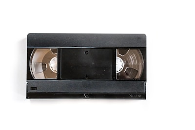 A black VHS tape lies on a white background. The central part features a rectangle with rounded corners, flanked by two open reels showing the tape spools inside.