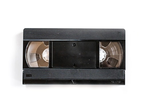 A black VHS tape lies on a white background. The central part features a rectangle with rounded corners, flanked by two open reels showing the tape spools inside.