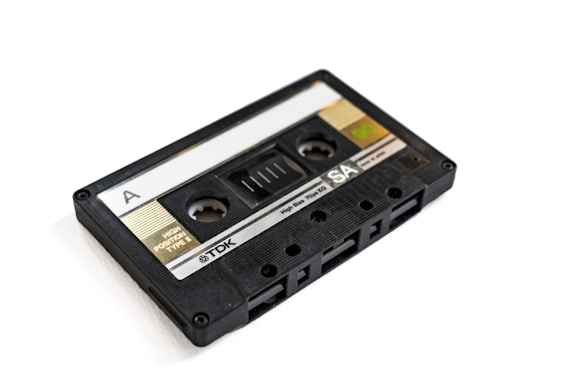 A vintage cassette tape with a black plastic casing and a transparent window, labeled with 'A' on one side and branding information including 'TDK' and 'SA'. The tape has visible spools and labeling indicating 'High Position Type II' and other technical specifications.