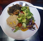 A white plate contains a serving of vegetable stir-fry, a portion of brown rice mixed with corn, and a fresh salad made of cucumbers, cherry tomatoes, and mixed greens. A fork rests on the edge of the plate.