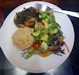 A white plate contains a serving of vegetable stir-fry, a portion of brown rice mixed with corn, and a fresh salad made of cucumbers, cherry tomatoes, and mixed greens. A fork rests on the edge of the plate.