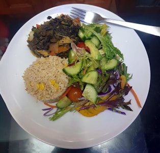 A white plate contains a serving of vegetable stir-fry, a portion of brown rice mixed with corn, and a fresh salad made of cucumbers, cherry tomatoes, and mixed greens. A fork rests on the edge of the plate.