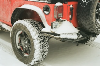 A red off-road vehicle is partially covered in snow, parked on a snowy surface. The rear tire and bumper are visible and also have snow accumulation, indicating cold weather conditions.