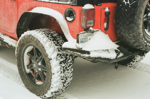 A red off-road vehicle is partially covered in snow, parked on a snowy surface. The rear tire and bumper are visible and also have snow accumulation, indicating cold weather conditions.