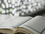 An open book written in Arabic script lies on a flat surface. It appears to be a religious or literary text, with intricate designs along the margins. The background features an out-of-focus decorative pattern that contrasts with the clear text.
