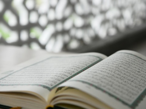 An open book written in Arabic script lies on a flat surface. It appears to be a religious or literary text, with intricate designs along the margins. The background features an out-of-focus decorative pattern that contrasts with the clear text.