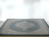 A close-up view of a book with a decorative and intricate cover design, featuring Arabic script, resting on a flat surface. The focus is on the cover's texture and pattern, while the background is softly blurred, giving a sense of calm and introspection.