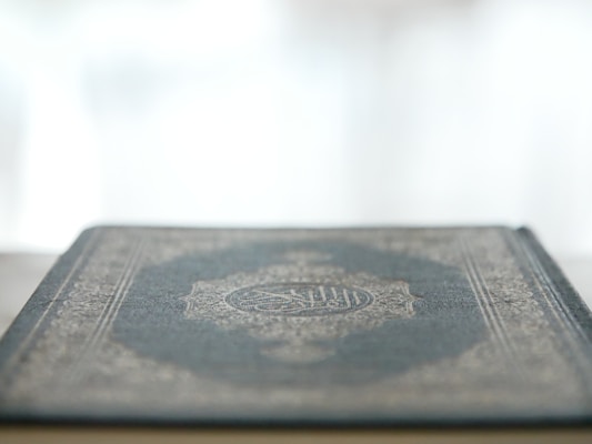 A close-up view of a book with a decorative and intricate cover design, featuring Arabic script, resting on a flat surface. The focus is on the cover's texture and pattern, while the background is softly blurred, giving a sense of calm and introspection.