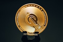 A close-up view of a gold-colored cryptocurrency token displaying intricate embossed designs and text around its circumference. The coin is positioned upright on a dark stand against a black background, highlighting its metallic sheen.