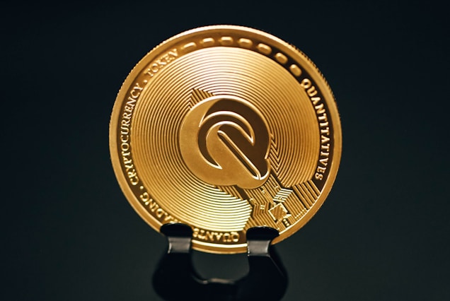A close-up view of a gold-colored cryptocurrency token displaying intricate embossed designs and text around its circumference. The coin is positioned upright on a dark stand against a black background, highlighting its metallic sheen.