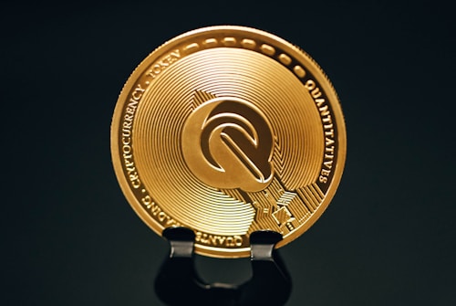 A close-up view of a gold-colored cryptocurrency token displaying intricate embossed designs and text around its circumference. The coin is positioned upright on a dark stand against a black background, highlighting its metallic sheen.