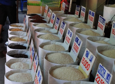 Numerous large bags of rice are displayed in a market setting, each labeled with a price and type in Vietnamese. The bags show different varieties of rice, varying in color from black to white. The photo conveys a traditional market atmosphere with the rice bags neatly arranged in rows.
