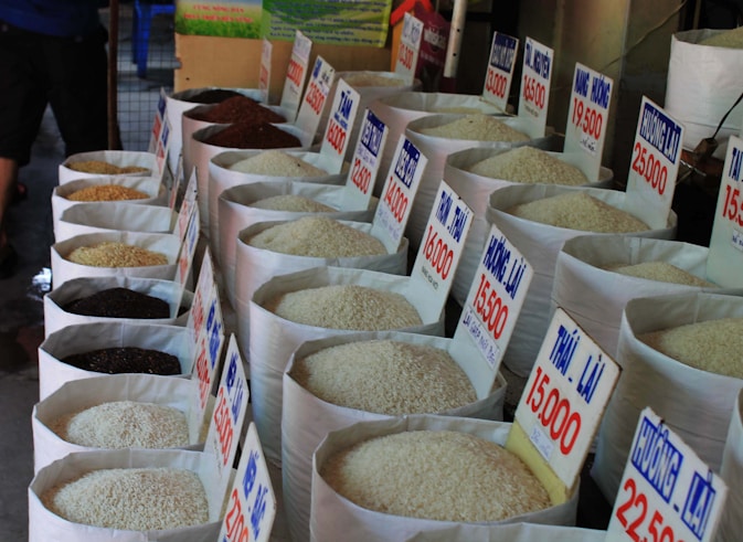 Numerous large bags of rice are displayed in a market setting, each labeled with a price and type in Vietnamese. The bags show different varieties of rice, varying in color from black to white. The photo conveys a traditional market atmosphere with the rice bags neatly arranged in rows.
