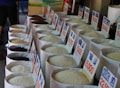 Numerous large bags of rice are displayed in a market setting, each labeled with a price and type in Vietnamese. The bags show different varieties of rice, varying in color from black to white. The photo conveys a traditional market atmosphere with the rice bags neatly arranged in rows.