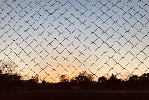 A chain-link fence in the foreground with a sunset in the background. The sky transitions from a light blue at the top to warm orange near the horizon. Silhouettes of trees and a few structures are visible against the evening sky.