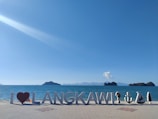 Large letters spell out 'I ♥ LANGKAWI' on a tiled promenade by the ocean, with a bright blue sky above. Several small decorative sculptures are positioned near the letters, including shapes resembling an anchor, a boat, and a tree. Distant islands and a lone cloud appear on the horizon.