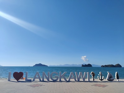 Large letters spell out 'I ♥ LANGKAWI' on a tiled promenade by the ocean, with a bright blue sky above. Several small decorative sculptures are positioned near the letters, including shapes resembling an anchor, a boat, and a tree. Distant islands and a lone cloud appear on the horizon.