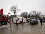 A street scene with several people holding signs and Canadian flags. There are vehicles, including a truck and a trailer, parked along the road. Trees with bare branches line the sides, indicating a cold season.