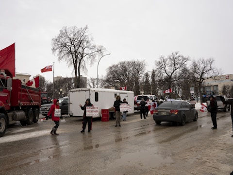 A street scene with several people holding signs and Canadian flags. There are vehicles, including a truck and a trailer, parked along the road. Trees with bare branches line the sides, indicating a cold season.