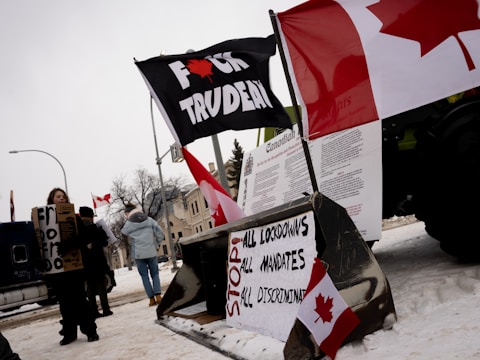 Protest scene with Canadian flags and signs displaying anti-government messages. The setting appears to be snowy, with several individuals holding placards. Prominent signs include phrases advocating for the end of lockdowns and mandates.