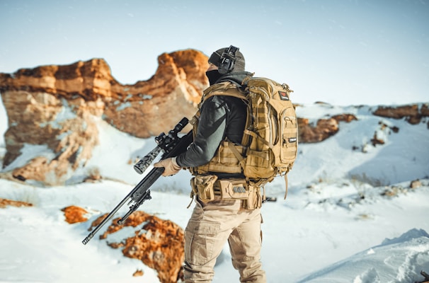 An individual wearing tactical gear and a large backpack stands in a snowy landscape. They are holding a rifle and appear to be facing scenic, sunlit rocks in the distance, with bright blue skies above.