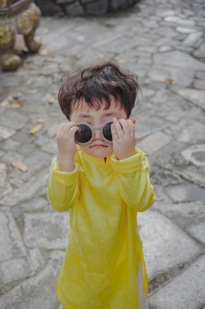 A young child in a bright yellow traditional outfit playfully holds large round sunglasses over their eyes. The child stands on a stone pathway, with scattered leaves around.