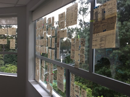 Numerous sheets of paper with diagrams and notes are attached to large glass windows. Outside the windows, lush green trees are visible, creating a backdrop for the working space. The papers appear to have sketches, charts, and written content, showcasing an organized thought or planning process.
