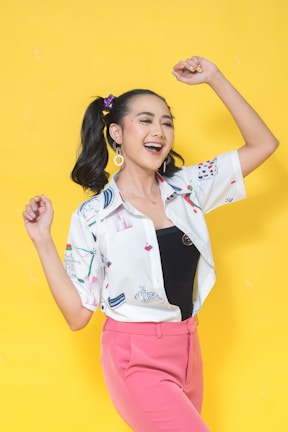 A joyful person is posing against a bright yellow background, wearing a patterned white shirt over a black top and pink pants. They have their hair styled in pigtails with colorful accessories and are smiling with arms raised.