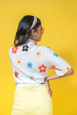 A person with long dark hair wearing a colorful floral shirt and light yellow pants stands against a solid yellow background. The shirt features bright, playful flower patterns in various colors. Large orange-tinted sunglasses and a headband complete the look, adding a retro style.