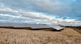 Rows of solar panels are installed on a grassy field under a partly cloudy sky. The landscape is open, with no trees in close proximity to the panels, allowing for maximum sun exposure. The grass appears brownish, indicating a dry season or late autumn.