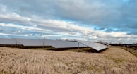 Rows of solar panels are installed on a grassy field under a partly cloudy sky. The landscape is open, with no trees in close proximity to the panels, allowing for maximum sun exposure. The grass appears brownish, indicating a dry season or late autumn.