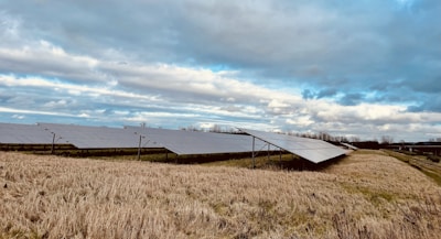 Rows of solar panels are installed on a grassy field under a partly cloudy sky. The landscape is open, with no trees in close proximity to the panels, allowing for maximum sun exposure. The grass appears brownish, indicating a dry season or late autumn.